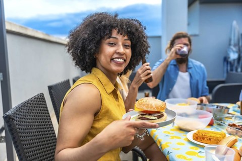 Young woman showing her delicious burger, enjoying a rooftop barbecue party with friends, drinking