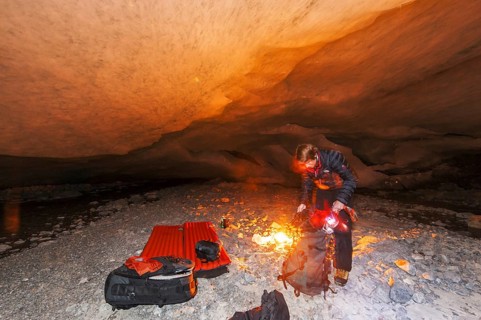 A person kneels by a campfire in a glacier cave, surrounded by darkness and warm light
