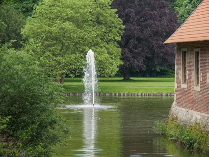 Fountain in a green park next to a brick building, with large trees and calm water, havixbeck,