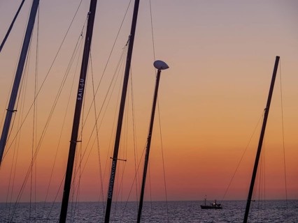 Masts in the foreground and a small boat in the sea at sunset, the sky is bathed in orange tones,