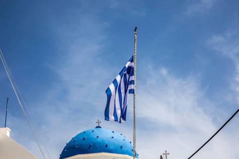Greek flag waving in the wind, blue church dome, Timios Stavros church, Perissa, Santorini island,