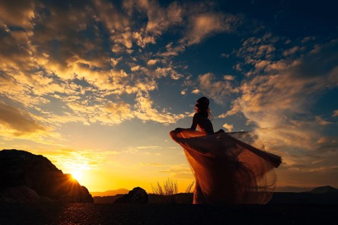 Bride in a fluttering dress stands on a mountain against the backdrop of a sunset sky.
