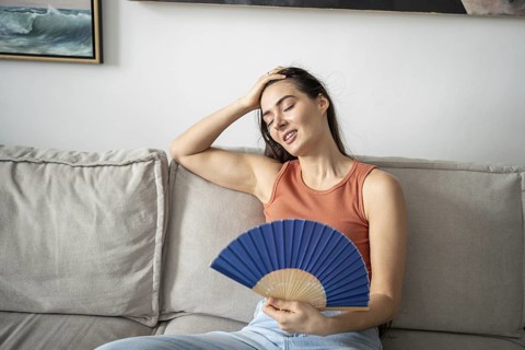 Satisfied woman smiling, blowing with fan, sitting on sofa in home studio. Happy pleased girl