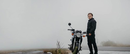 A young man stands by a motorcycle on a foggy road, looking ahead