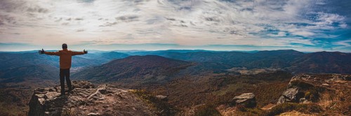 Panoramic view of mountain valley. Young man standing with hands widespread trying to embrace whole