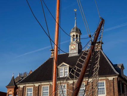 Building with clock tower and sailing mast in the foreground under a blue sky, empty, East Frisia,