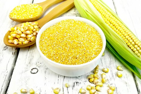 Corn grits in a white bowl and spoon, cobs and grains on the background of wooden boards
