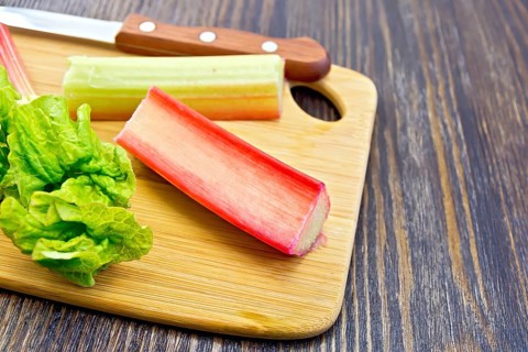 Rhubarb stalks with green leaves and a knife on a small planch on a wooden boards background