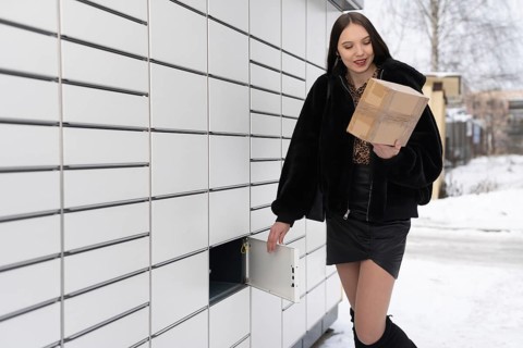 An attractive student collects and sends a parcel via a parcel locker in winter. Automatic machine