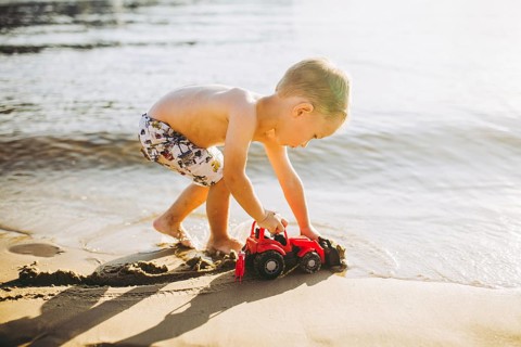 Caucasian child boy plays toy red tractor, excavator on sandy beach by the river in shorts at