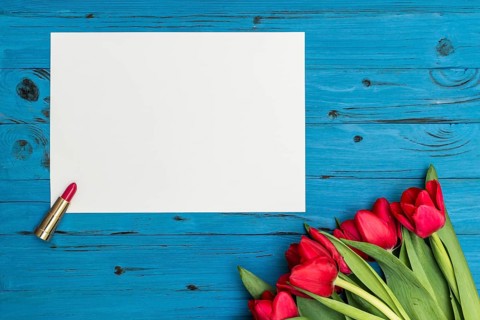 Red tulips, lipstick and a white sheet of paper for your greetings on the background of blue wooden