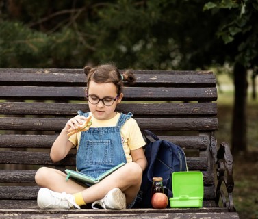 Cute little schoolgirl eating from lunch box outdoor sbtting on a bench. Food for kids