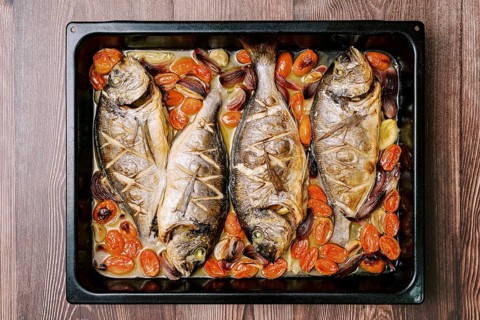 Baked sea bream with vegetables on a baking sheet stands on a wooden table. Top view.