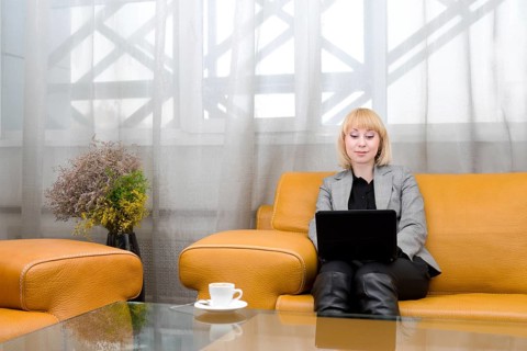 Young blonde woman sits on leather sofa with laptop, in front of glass table