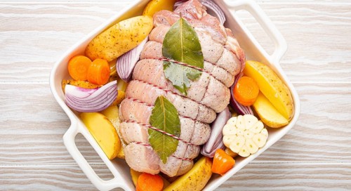 Raw wrapped rolled sliced pork in white casserole dish with potatoes, vegetables and herbs on