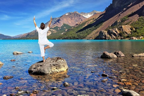 Yoga on the lake. Mature woman in white performs asana tree, standing on a rock