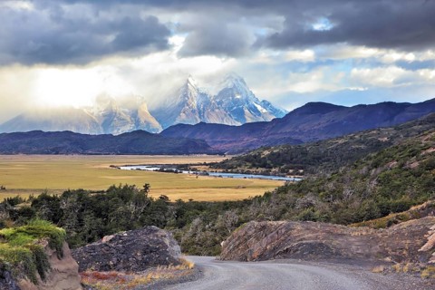 The road between turned yellow hills and river goes to snow-covered black rocks. Patagonia.
