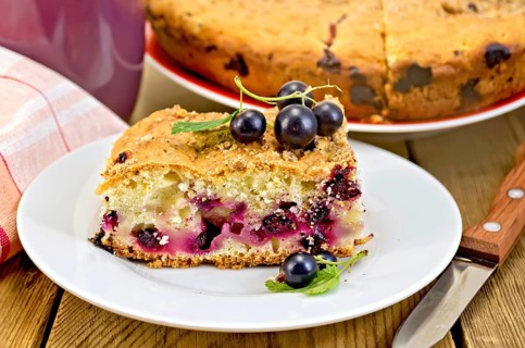 Sweet piece of cake with black currant, napkin, cup, knife on background wooden plank