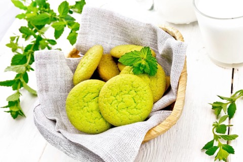Green mint cookies on napkin in wicker basket, milk in a glass and a jug on white wooden board