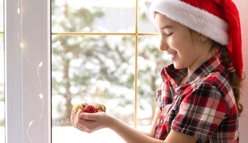 Girl in a Santa hat sits on the windowsill of a house near the Christmas tree and holding glass