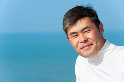 Handsome asian young man looks into camera smiling, blue sea and sky
