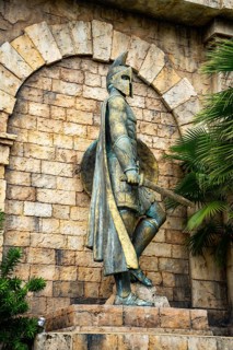 Close-up of an ancient statue with helmet and shield amidst stonework and greenery, palms, city,