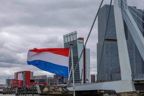 Dutch flag waving in front of a bridge and modern buildings under a cloudy sky, Rotterdam,
