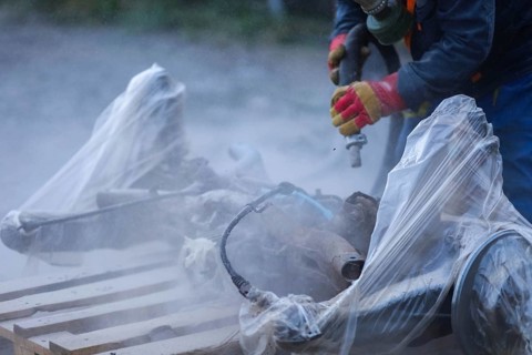 A worker in protective gloves sanding car parts covered in plastic, producing dust