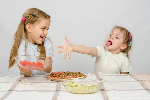 Two little girls at a table prepared pizza. One with a whimsical view stretches a hand to a plate