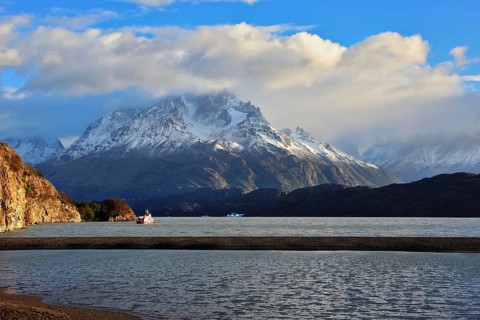 Gray lake and snow-capped mountains. By the banks of the red tourist ship sails. Summer sunset