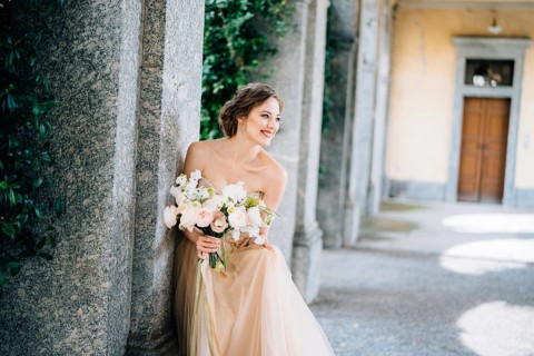 Smiling bride in a dress with a bouquet of pink flowers sat down near a pillar in a vaulted room.