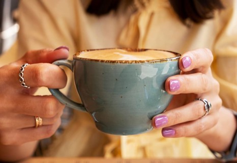 Woman in yellow clothes holding blue cup of cappuccino with both hands, close-up