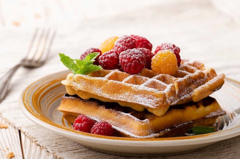 Belgian waffles served with raspberries and mint leaf dusted with powdered sugar on white wooden