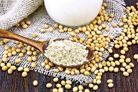 Soy flour in a spoon and milk in glass jug, soybeans on sackcloth on wooden board background