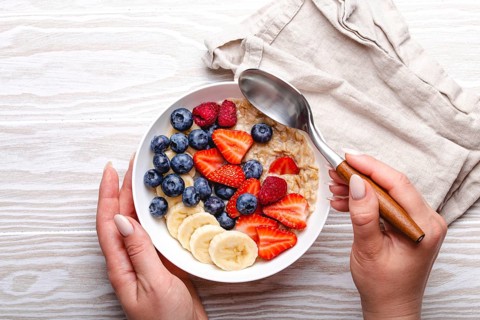Top view of female hands holding bowl with oatmeal porridge with fruit and berries and spoon on