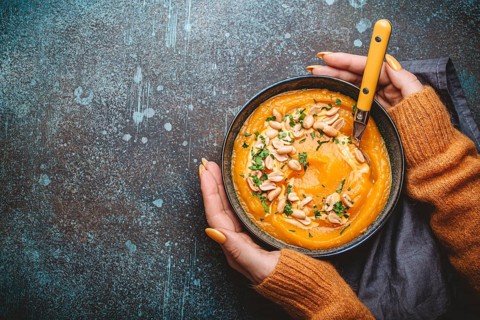 Female hands in yellow knitted sweater holding a bowl with pumpkin cream soup on dark stone