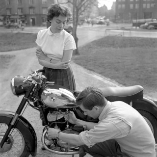 In the 1950s. A man on his Puch motorcycle with a woman looking at him while he checks the fuel