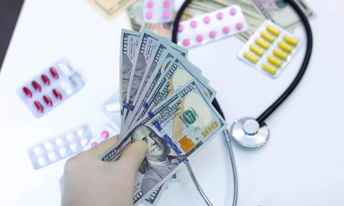 Hand with cash, stethoscope, and pills, illustrating high cost of healthcare