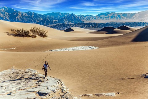 Elderly woman with a camera and a tripod goes among the dunes. Light sand waves from the desert
