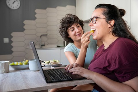 A happy lesbian couple enjoys feeding each other while working. Their laughter, casual attire, and