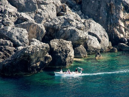 A small boat sails along the rocky coast in clear blue water, rocky blue sea with green bushes and