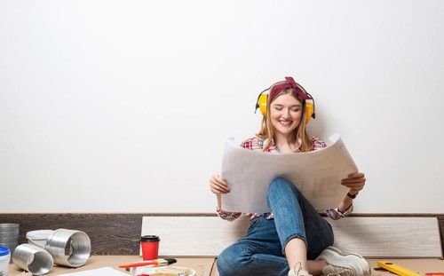 Happy girl in protective headphones sitting on floor with paper blueprint. Home remodeling after
