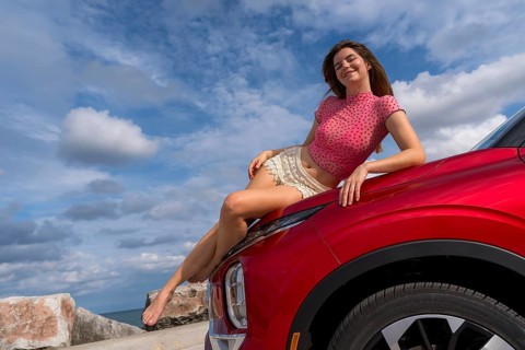 A gorgeous redhead model sits confidently on the hood of her car, soaking in the summer breeze as