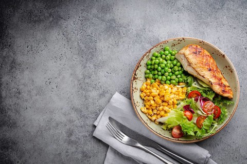 Baked chicken breast on plate with fresh salad, green peas and corn, grey stone background, top