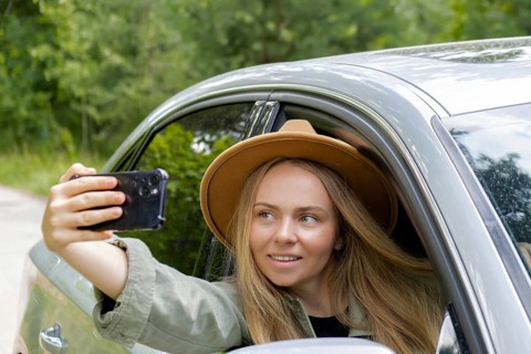 Exited smiling woman making video call with mobile phone from car window. Local solo travel on