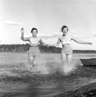 1950s summer. Two young women runs on the shallow part of the beach and the water splashes. Sweden