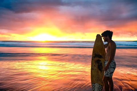 A surfer stands with surfboard in hand on the beach and watches the sun rise over the ocean on a