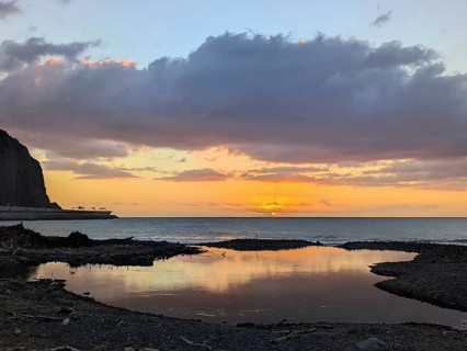 Sunset over the ocean, with striking reflections on wet sand and dramatic clouds in the sky.