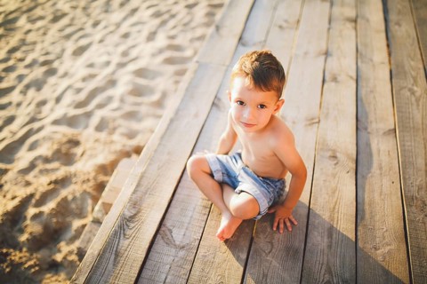 Little Caucasian boy child sitting on wooden pier sandy beach, summer time, sea holiday near water.