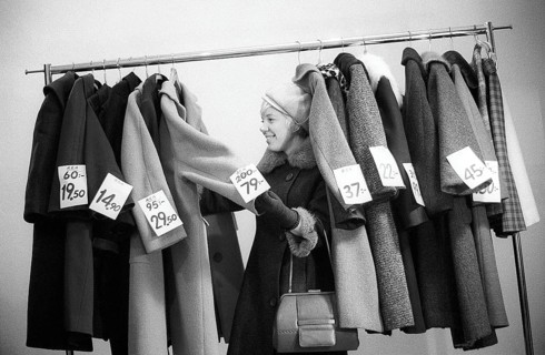 Great discounts of outerwear in the 1960s. A woman stands by a clothes rack, choosing from